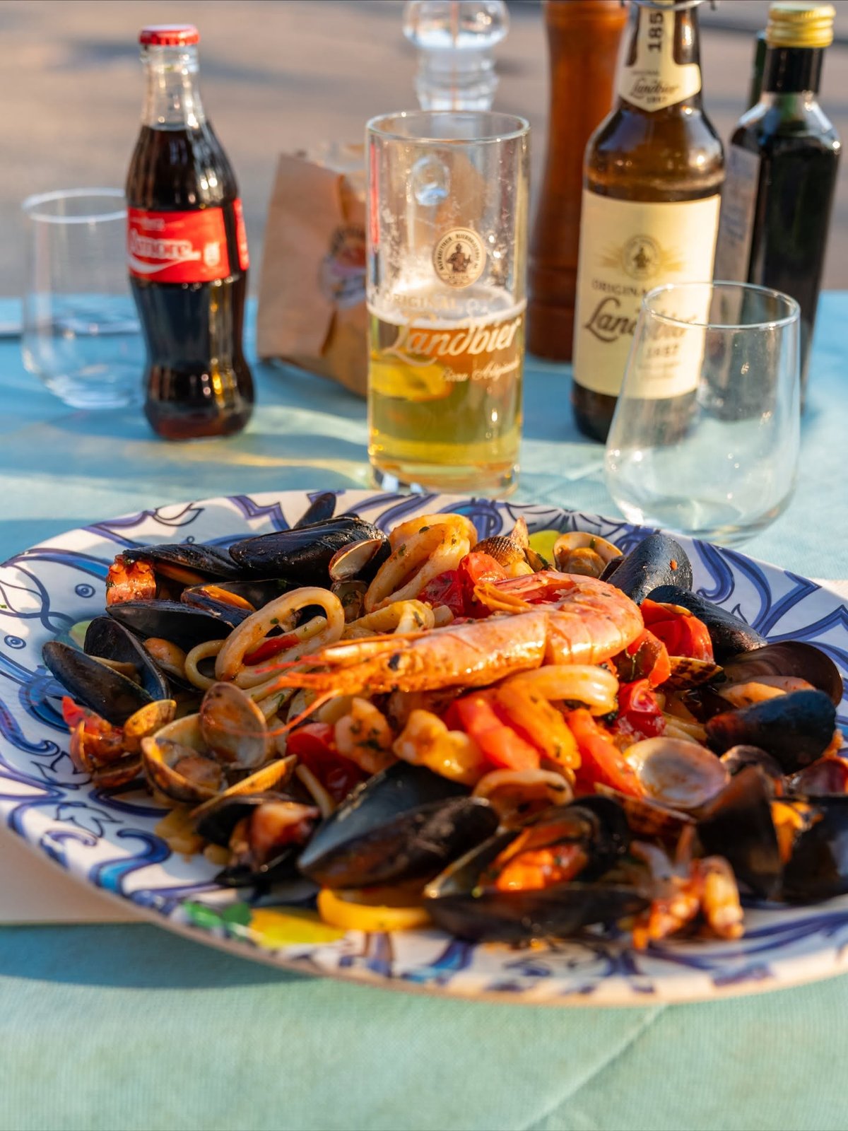 Piatto di frutti di mare con cozze, vongole, calamari e gambero su ceramica blu, birra Landbier e Coca-Cola sul tavolo