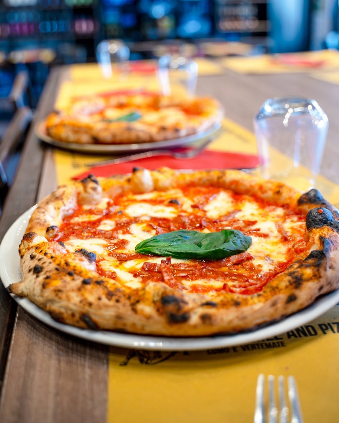 Pizza OX Como con salame piccante, mozzarella e basilico su tovaglietta gialla, dietro un'altra pizza e bicchieri