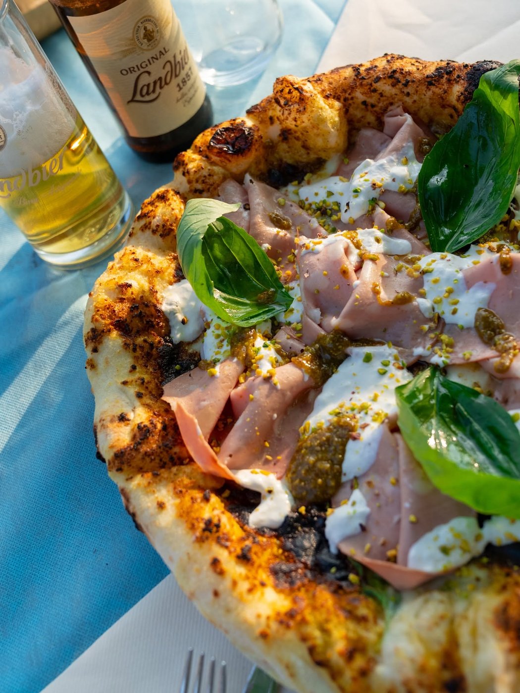 Pizza di OX Como con mortadella, burrata, pistacchio e basilico, accanto a birra Original Landbier su tovaglia azzurra